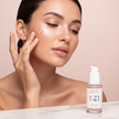Close-up of a woman with clear, glowing skin applying a skincare serum with droplets on her cheek, featuring a white and pink bottle of LUMINARA Skin Renaissance Serum on marble surface